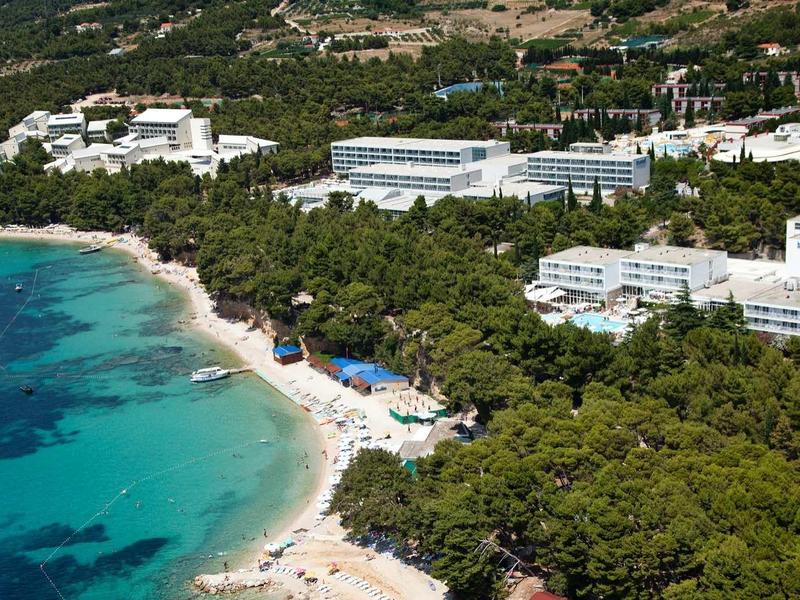 Küstenlinie mit Sandstrand, blauem Meer, grünen Pinienwäldern und mehreren weißen Gebäuden.