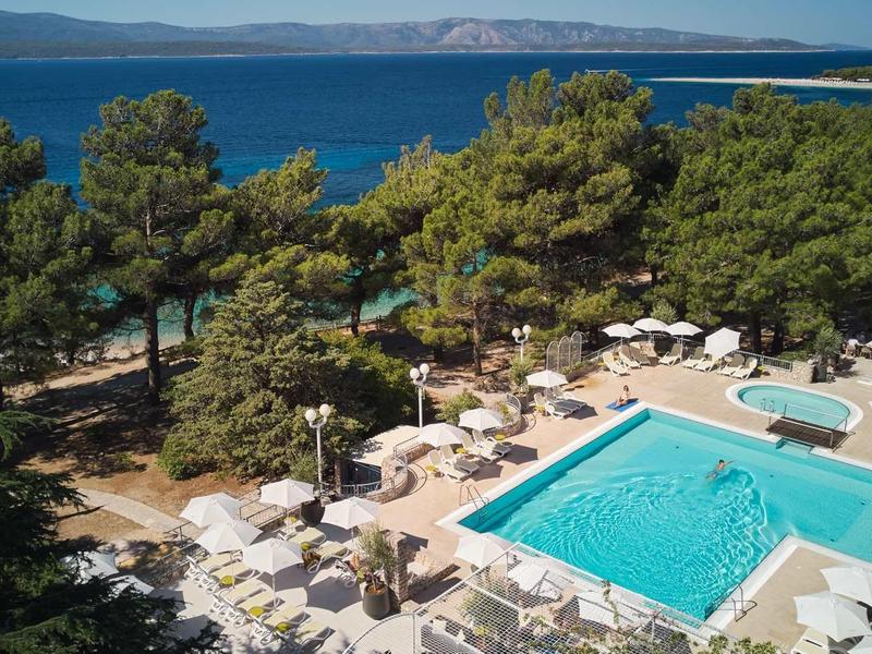 Freizeitbereich mit Pool, Sonnenschirmen und Liegen, umgeben von Bäumen und Meer im Hintergrund.