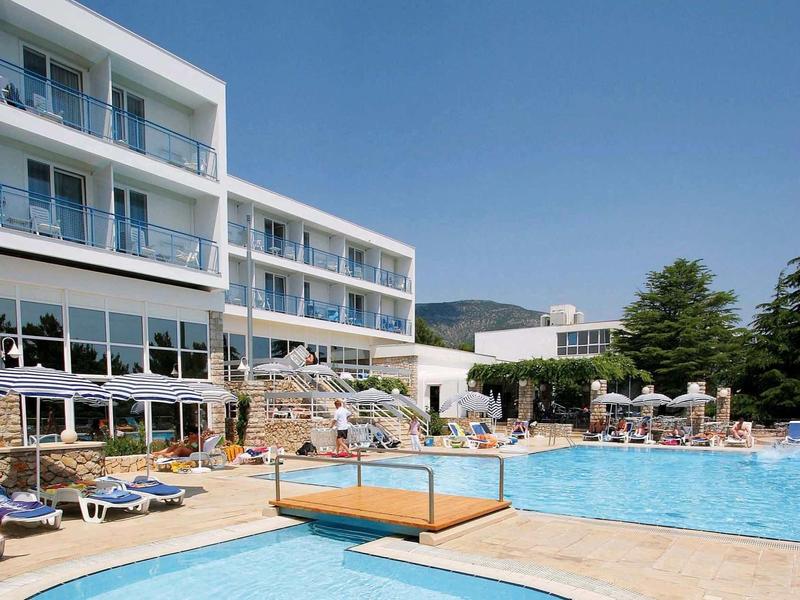 Großer Außenpool mit Sonnenterrasse, modernen Gebäuden und blauem Himmel im Hintergrund.