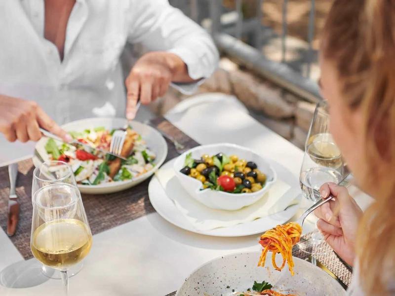 Zwei Personen essen draußen, Teller mit Salat und gefüllten Oliven, Gläser mit Getränken auf dem Tisch.