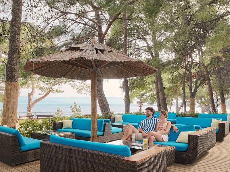 Outdoor-Lounge mit blauen Sofas, Sonnenschirm und grünen Bäumen vor einem hellen Himmel.