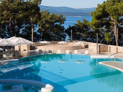 Poolbereich mit blauen Wasser, weißen Liegen und Blick auf das Meer hinter Bäumen.
