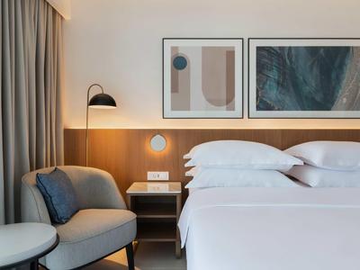 Modern hotel room with white bed, armchairs, bedside table, and abstract wall art.