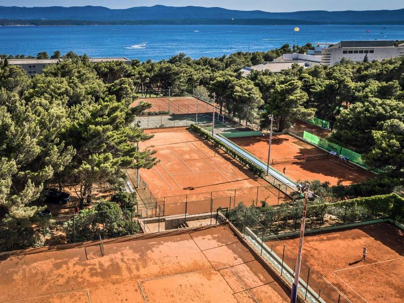 Several red clay tennis courts surrounded by trees with a sea view in the background.