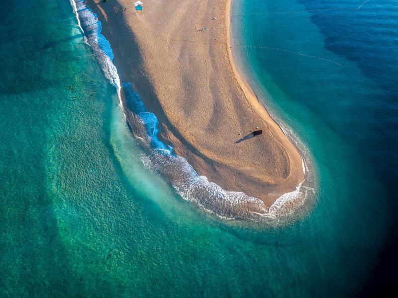 Aerial view of a sand spit extending into clear blue sea with gentle waves.