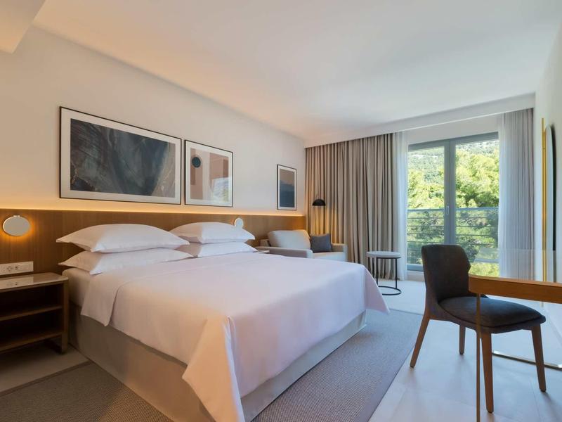 Modern hotel room with large bed, desk, chair, and view of green garden.