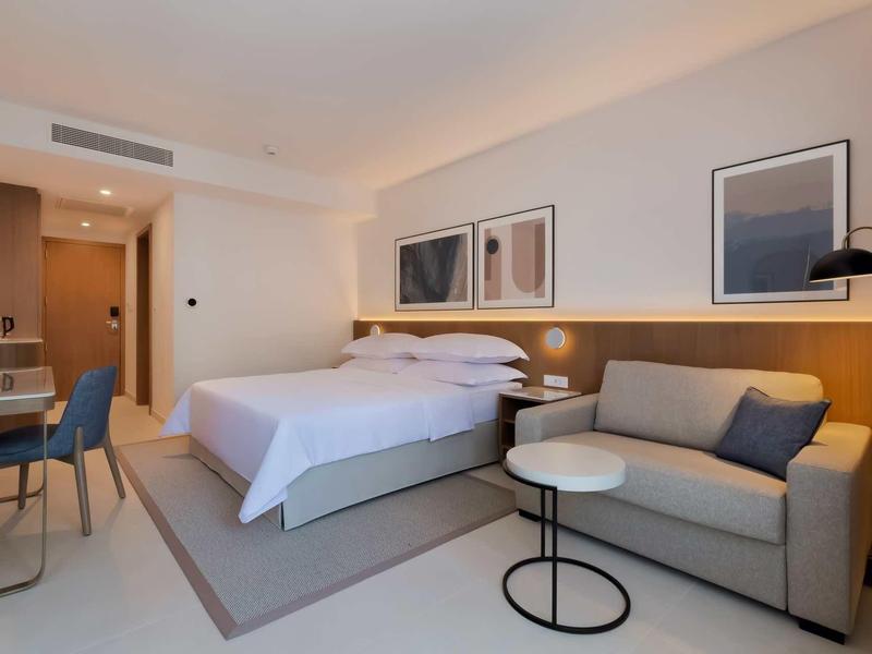 Modern hotel room with large bed, sofa, and desk in light tones.