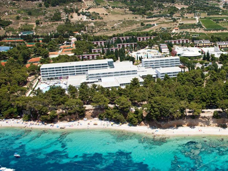 Large hotel complex by the beach with turquoise sea and wooded surroundings.