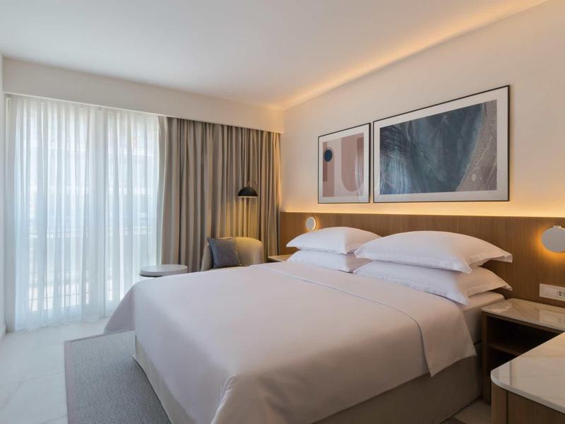 Bright hotel room with double bed, artwork on wall, and large windows with curtains.