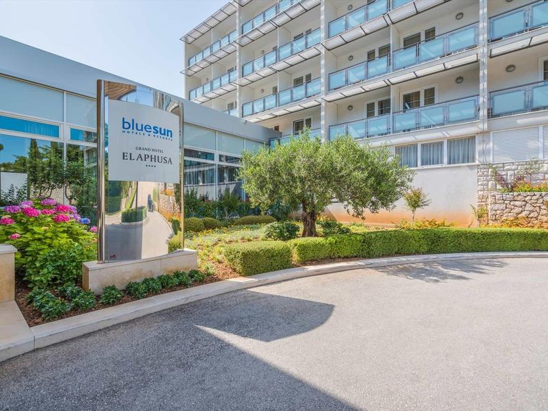 Modern hotel exterior with flower pots, bus stop, and large building in the background.