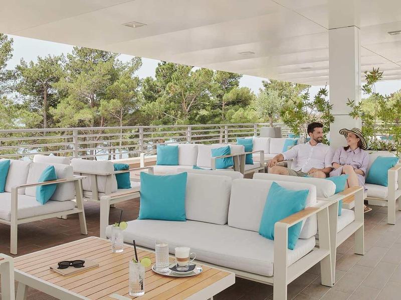 Modern hotel terrace with comfy sofas and turquoise cushions, guests are relaxing.