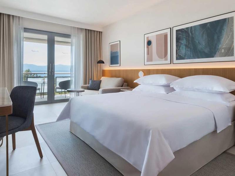 Bright hotel room with large bed, desk, and balcony with view.