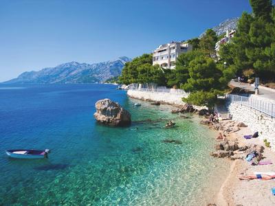 Clear water beach with rocks, boat, and tourists under blue sky and green trees.