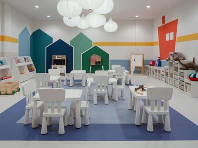 Children's playroom with white tables and chairs, colorful house wall decorations, and shelves.