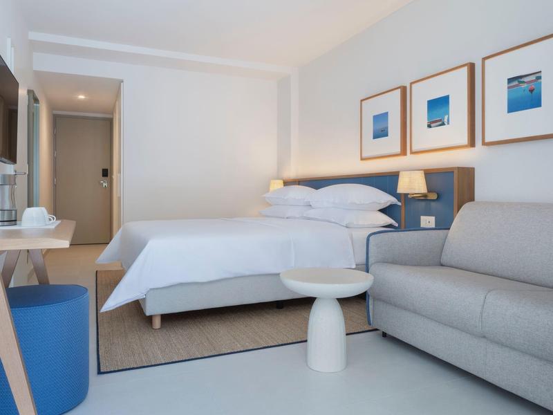 Modern hotel room with bed, sofa, TV, and framed artwork on the wall.