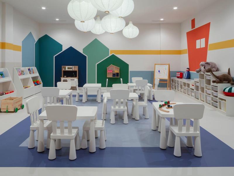 Children's playroom with white tables and chairs, colorful house wall decorations, and shelves.
