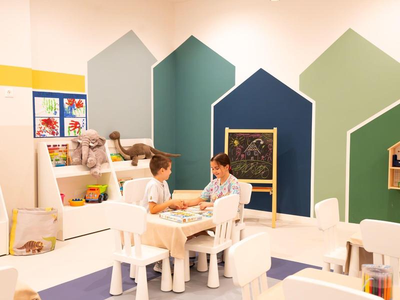 Two children sit at a table in a colorful playroom with house-shaped wall decorations.