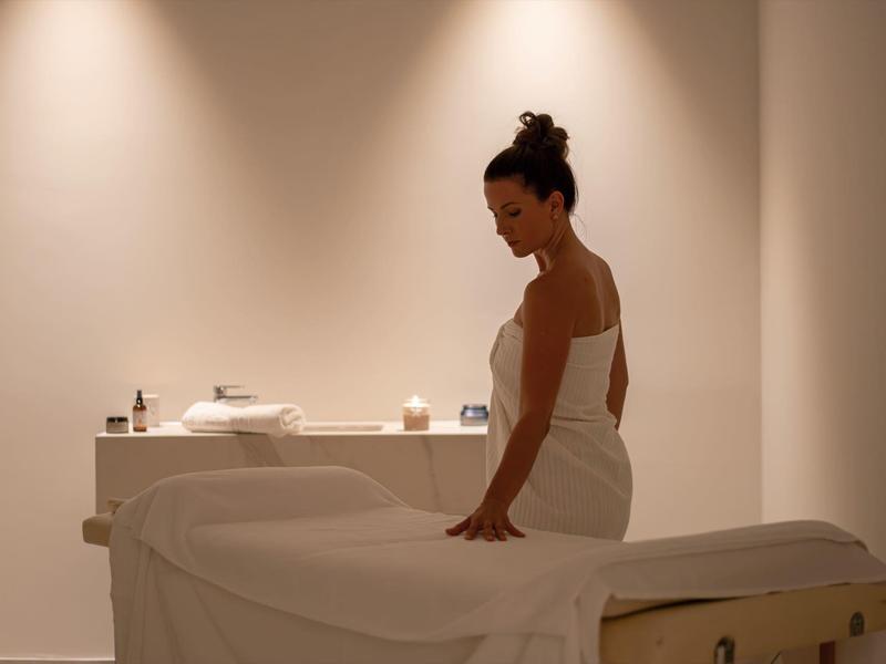 Woman in white towel attire sits on massage table in calm, stylish room.