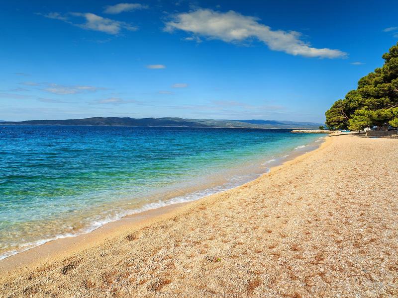 A calm sandy beach with clear blue water and forested hills in the background.