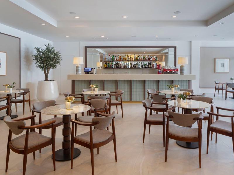 Bright, modern hotel bar area with wooden tables, armchairs, and decorative plants.