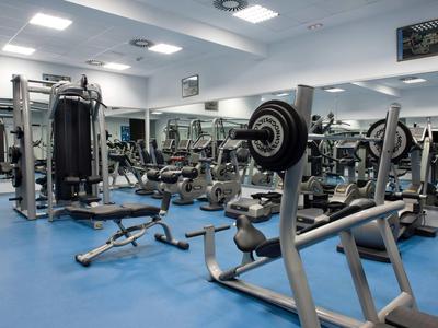 Fitnessraum mit verschiedenen Geräten und Spiegeln, blaue Bodenmatten und weiße Wände.