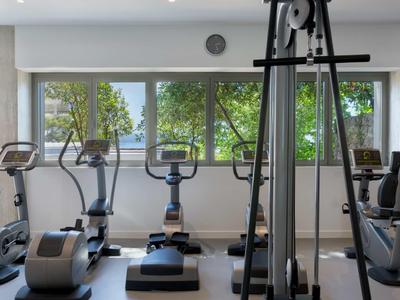 Fitnessraum mit verschiedenen Trainingsgeräten und großer Fensterfront mit Blick auf Bäume.