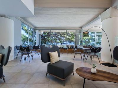 Heller, moderner Raum mit großen Fenstern, schwarzen Sesseln, Holztischen und hellem Fliesenboden.