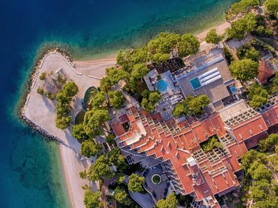 Luftaufnahme eines roten Gebäudekomplexes mit Pool, umgeben von Bäumen und blauem Wasser.