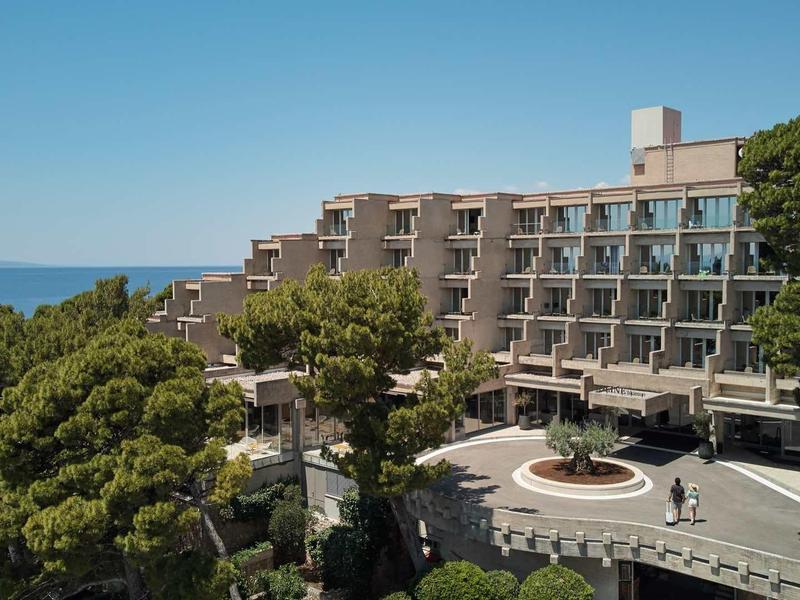 Großes mehrstöckiges Hotelgebäude mit Glasfassade, umgeben von grünen Bäumen und blauem Himmel.