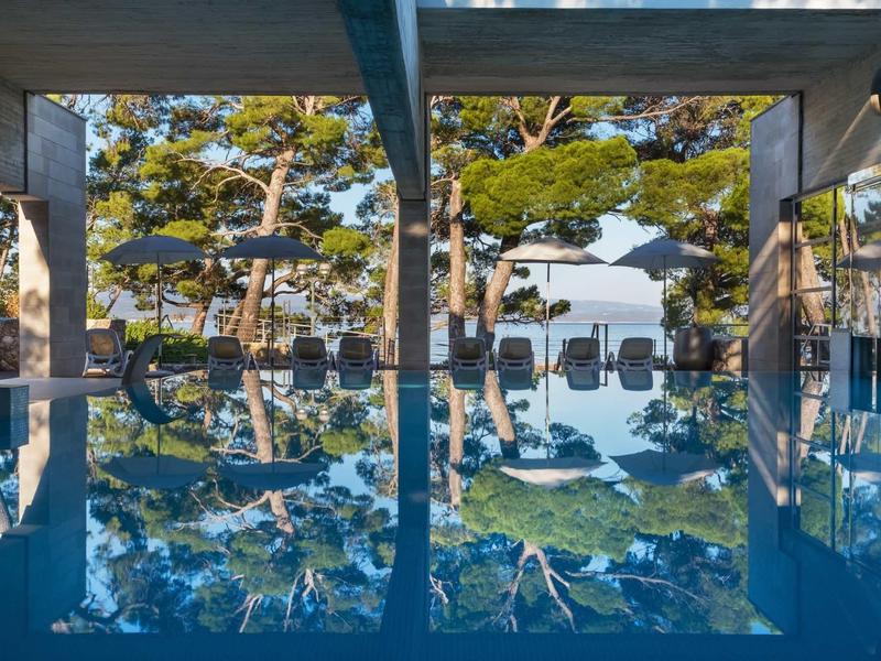 Ein blauer Pool spiegelt Bäume und Sonne, umgeben von Liegestühlen unter Betonbalken.