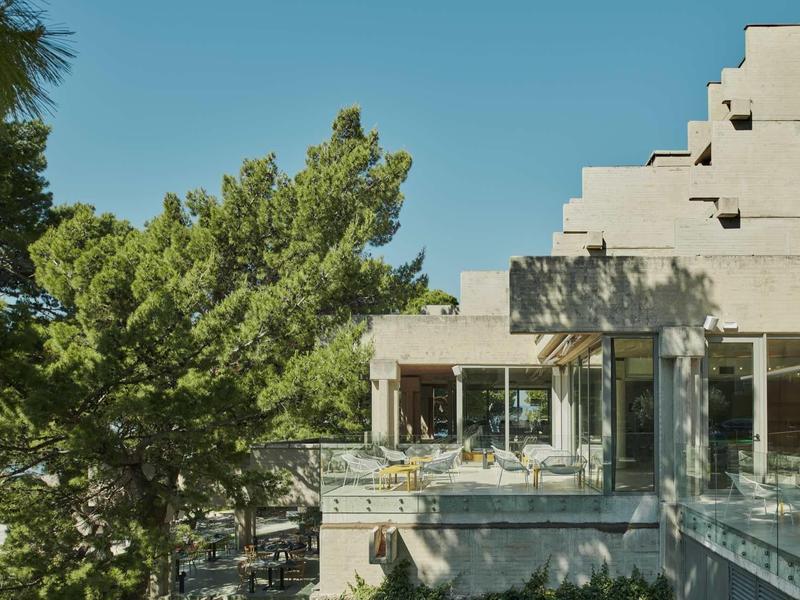 Modernes Gebäude mit großen Fenstern, umgeben von grünen Bäumen unter klarem blauem Himmel.