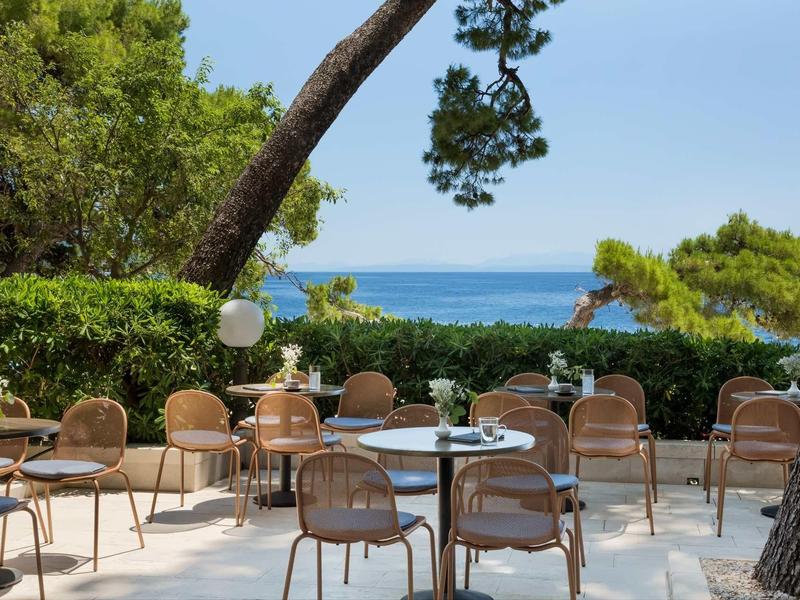 Leere Terrasse mit runden Tischen und Stühlen, umgeben von grünen Bäumen mit Meerblick.