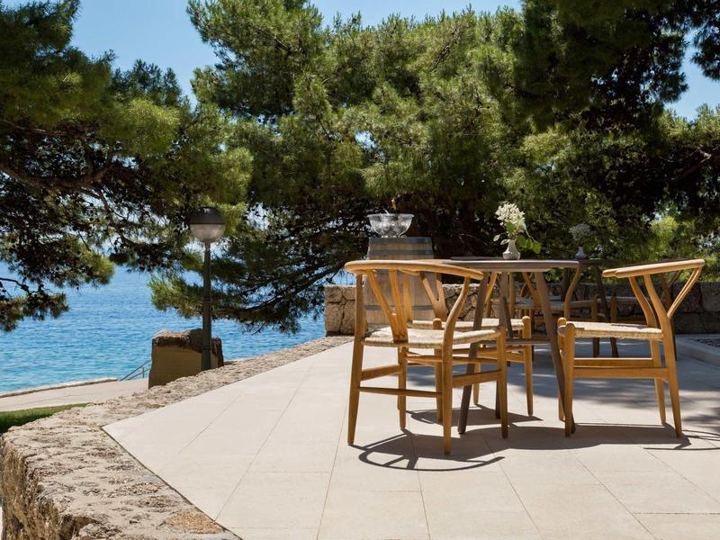 Terrasse mit Holzstühlen und Tisch, umgeben von Pinien, Blick aufs blaue Meer im Hintergrund.