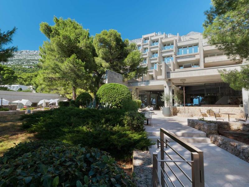 Modernes mehrstöckiges Gebäude umgeben von grünen Bäumen und Buschwerk unter blauem Himmel.