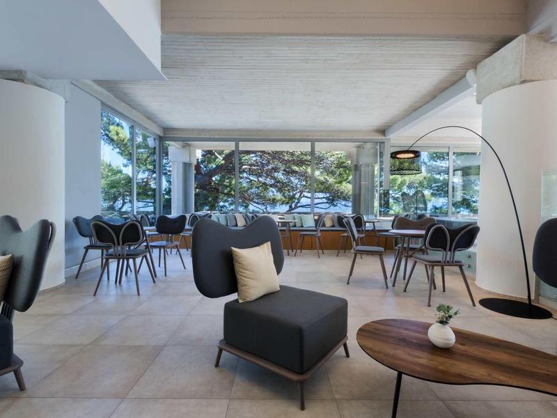 Heller, moderner Raum mit großen Fenstern, schwarzen Sesseln, Holztischen und hellem Fliesenboden.