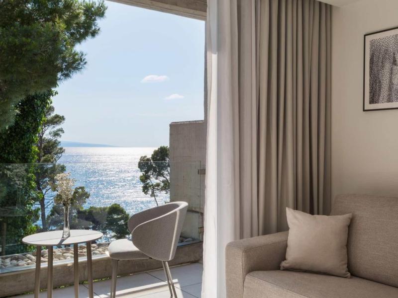 Helle Wohnung mit Couch, Kissen, Vorhängen und Balkon mit Stühlen, Tisch und Meerblick.