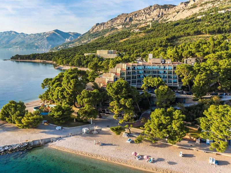 Küstenlandschaft mit Sandstrand, blauem Meer, grünen Bäumen und Bergen im Hintergrund.