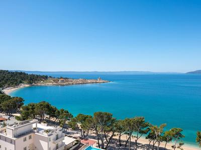 Küste mit blauem Meer, grün bewaldeten Hügeln und weißen Gebäuden im Vordergrund bei klarem Himmel.
