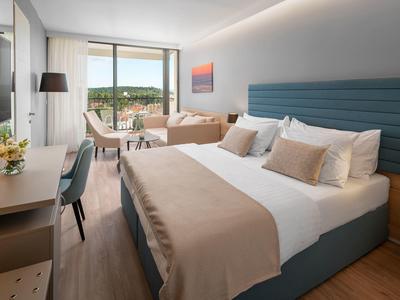 Modernes Hotelzimmer mit Doppelbett, Schreibtisch, Sessel und Balkon mit Aussicht auf Grünfläche.