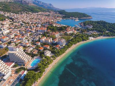 Städtische Küstenlandschaft mit Sandstrand, klarem Wasser, Gebäuden und bewaldeten Hügeln.