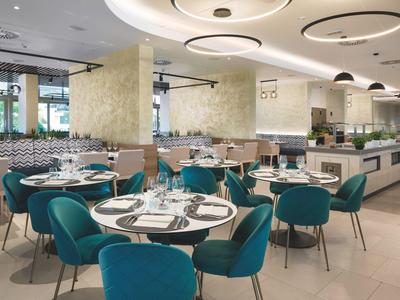 Moderner heller Restaurantbereich mit runden Tischen, blauen Stühlen und stilvoller Deckenbeleuchtung.