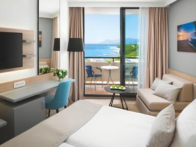 Modernes Hotelzimmer mit Doppelbett, Sofa, Schreibtisch, Balkon und Meerblick bei Tageslicht.