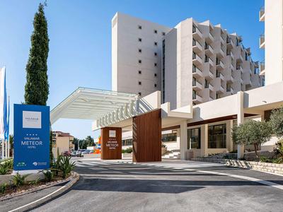 Moderner Hotelkomplex mit gepflegter Einfahrt, großen Glasüberdachungen und blauen Fahnen.