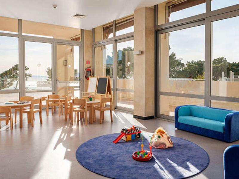 Heller Gruppenraum mit blauen Sitzmöbeln, Holzstühlen, Tischen und Spielzeug auf rundem Teppich.