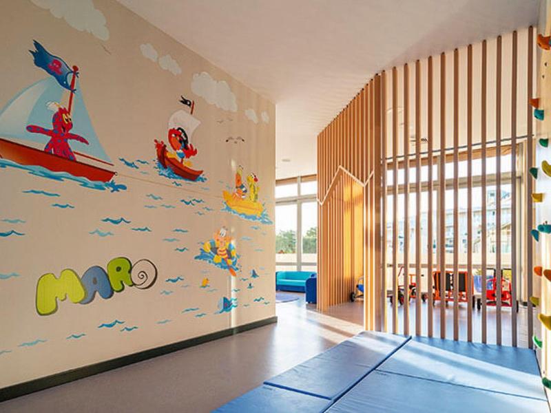 Heller Raum mit bunten Wandbildern und Kletterwand, trennend durch Holzlatten und blauen Bodenmatten.