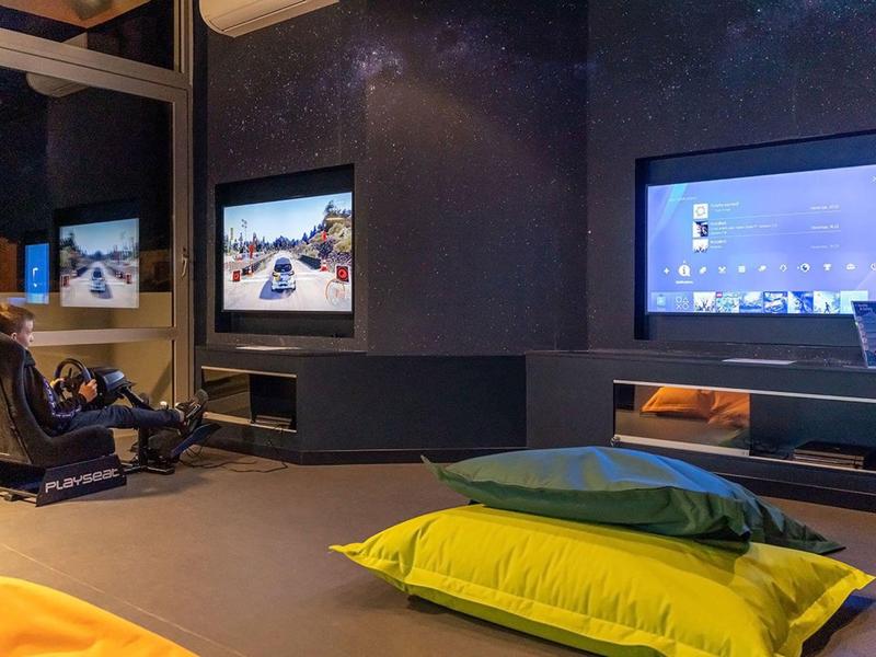 Moderner Raum mit großer Fensterfront, zwei Bildschirmen, Gaming-Stuhl und bunten Sitzkissen.