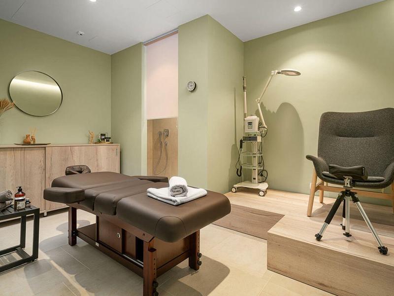 Moderner, heller Behandlungsraum mit Holzmöbeln, Massagebank, Rollstuhl und runden Spiegel an Wand.