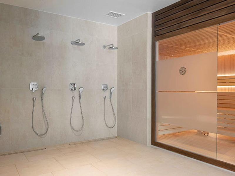 Moderne Duschanlage mit vier Duschen neben einer Sauna mit Holzwänden und Glasfront.