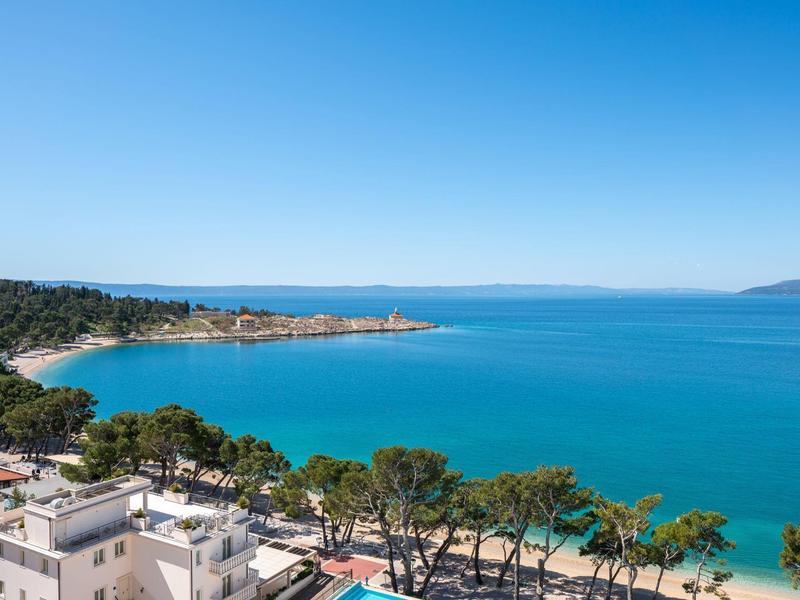 Küste mit blauem Meer, grün bewaldeten Hügeln und weißen Gebäuden im Vordergrund bei klarem Himmel.