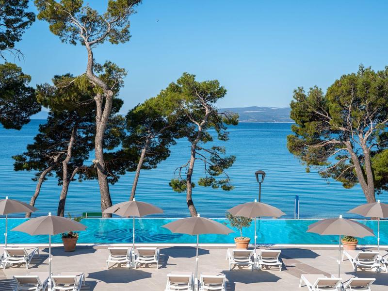 Poolbereich mit weißen Liegen, Sonnenschirmen und Bäumen vor ruhigem blauem Meer und klarem Himmel.